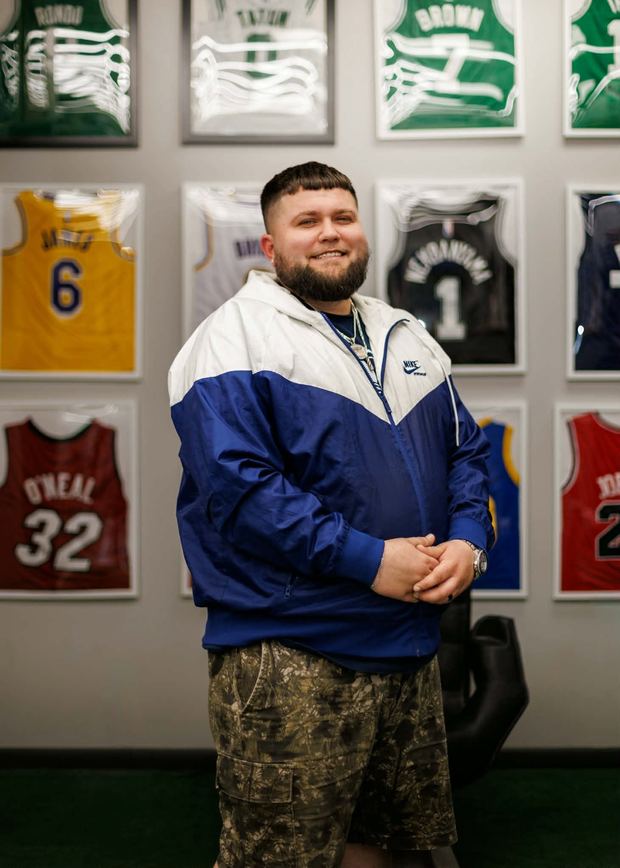 Bryndyn Lawrenz standing in a Railside windbreaker in front of a wall of framed basketball jerseys, hands clasped, smiling at the camera.