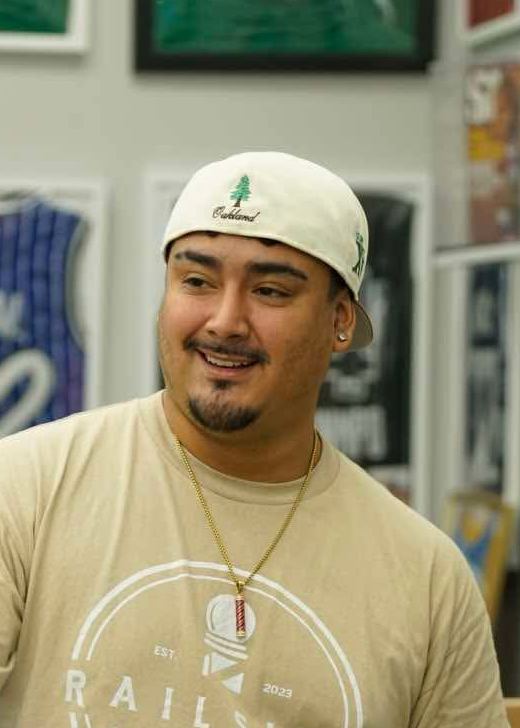 Isaiah Zapata at Railside in a white cap and tan Railside tee, smiling, in front of the shop's framed-jersey wall