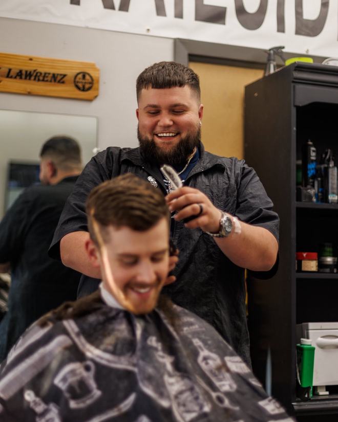 Bryndyn Lawrenz smiling as he runs the trimmers around a caped client's temple, his LAWRENZ station sign on the wall behind them.