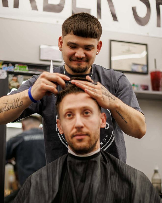 Shad Quintanilla combing and scissor-cutting the top of a caped client's hair, both of them facing the mirror.