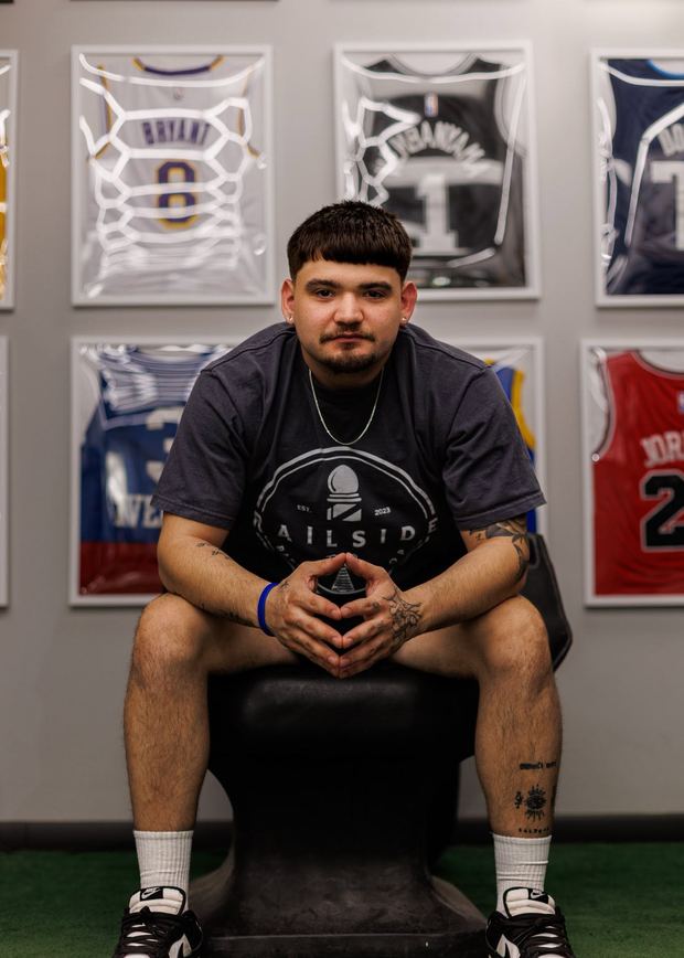 Shad Quintanilla seated on a black stool in a Railside tee, forearm tattoos showing, in front of the same framed-jersey wall.